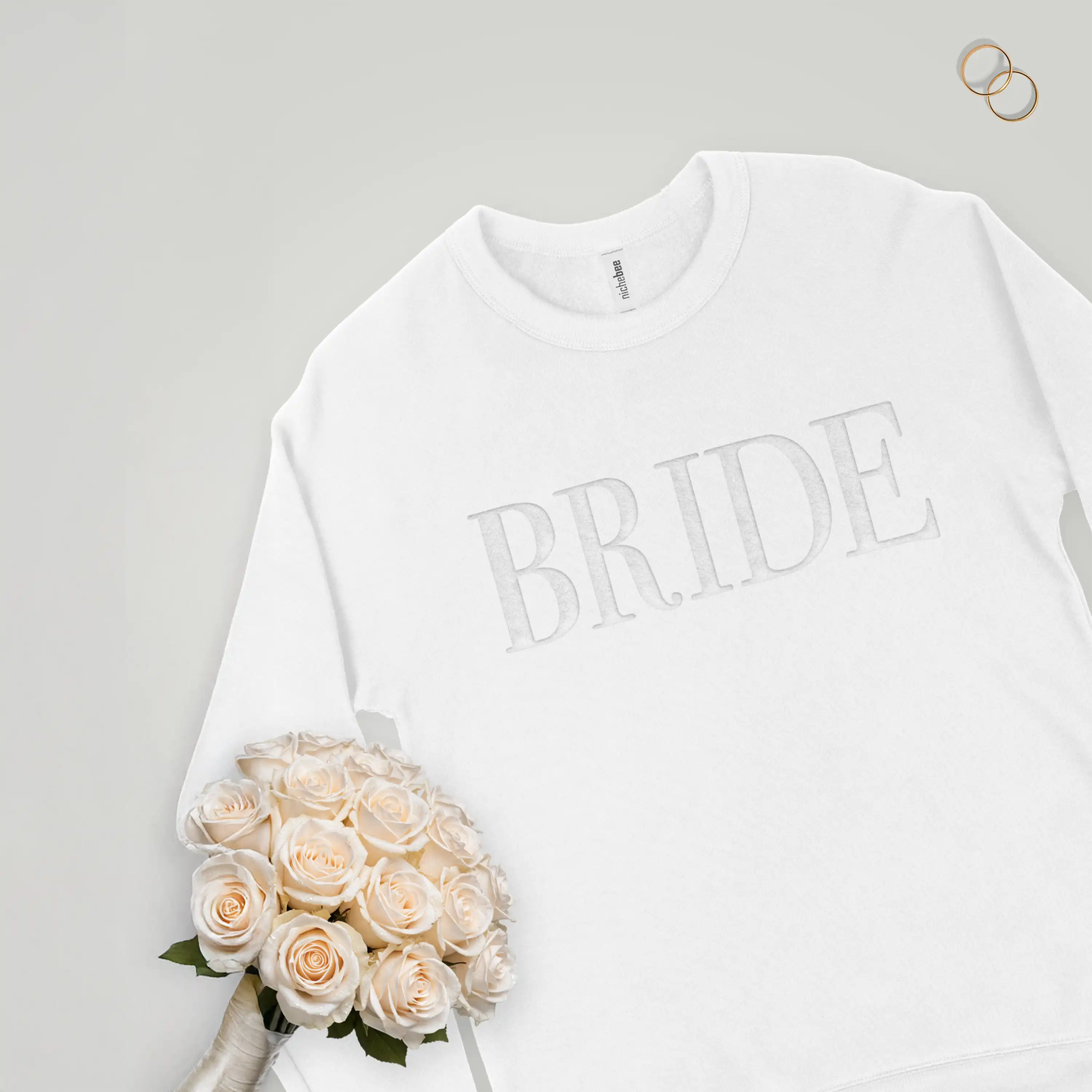 White sweatshirt with 'BRIDE' text, bouquet of white roses, and wedding rings on a light gray background