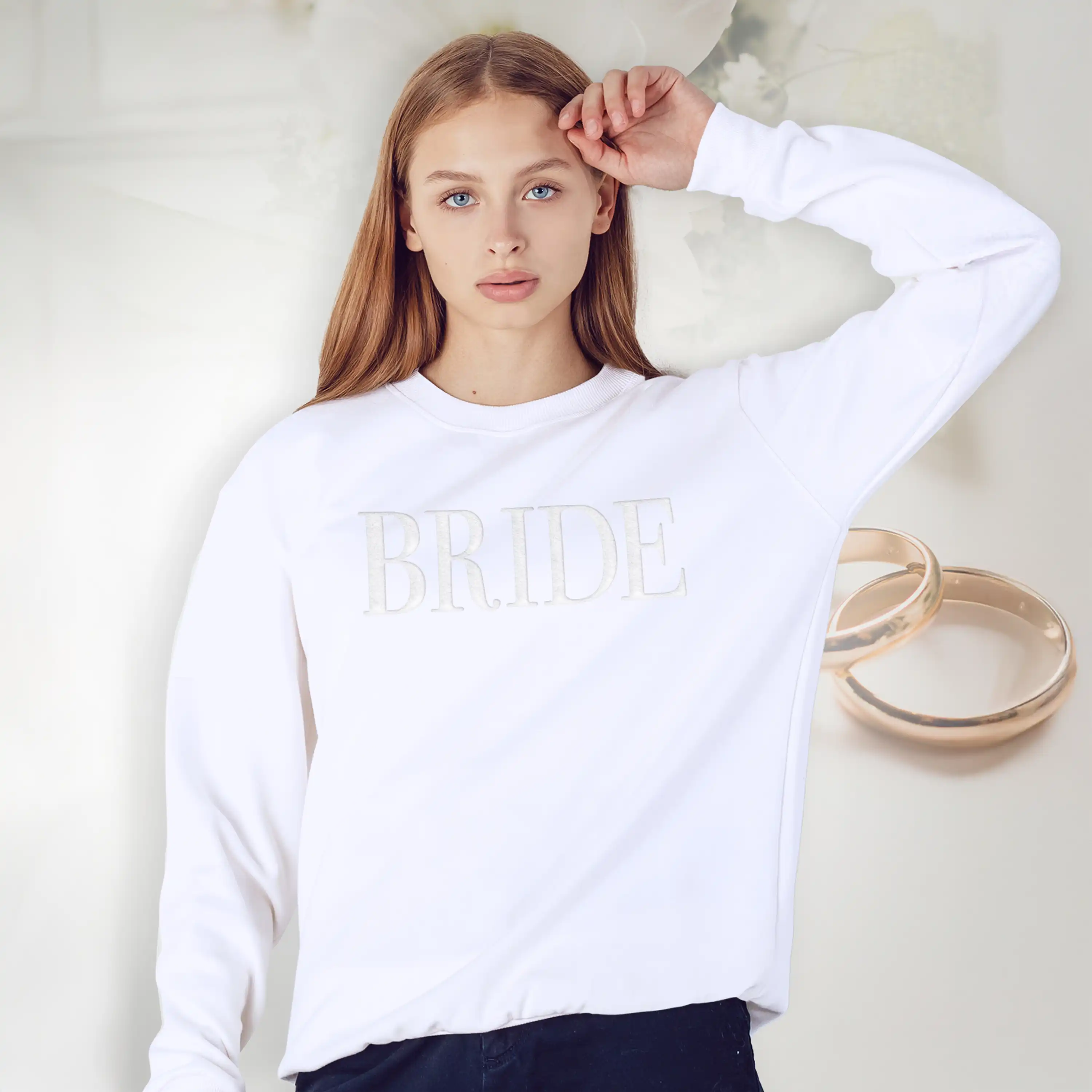 Woman wearing a white sweatshirt with 'BRIDE' text, standing next to gold rings on a light background
