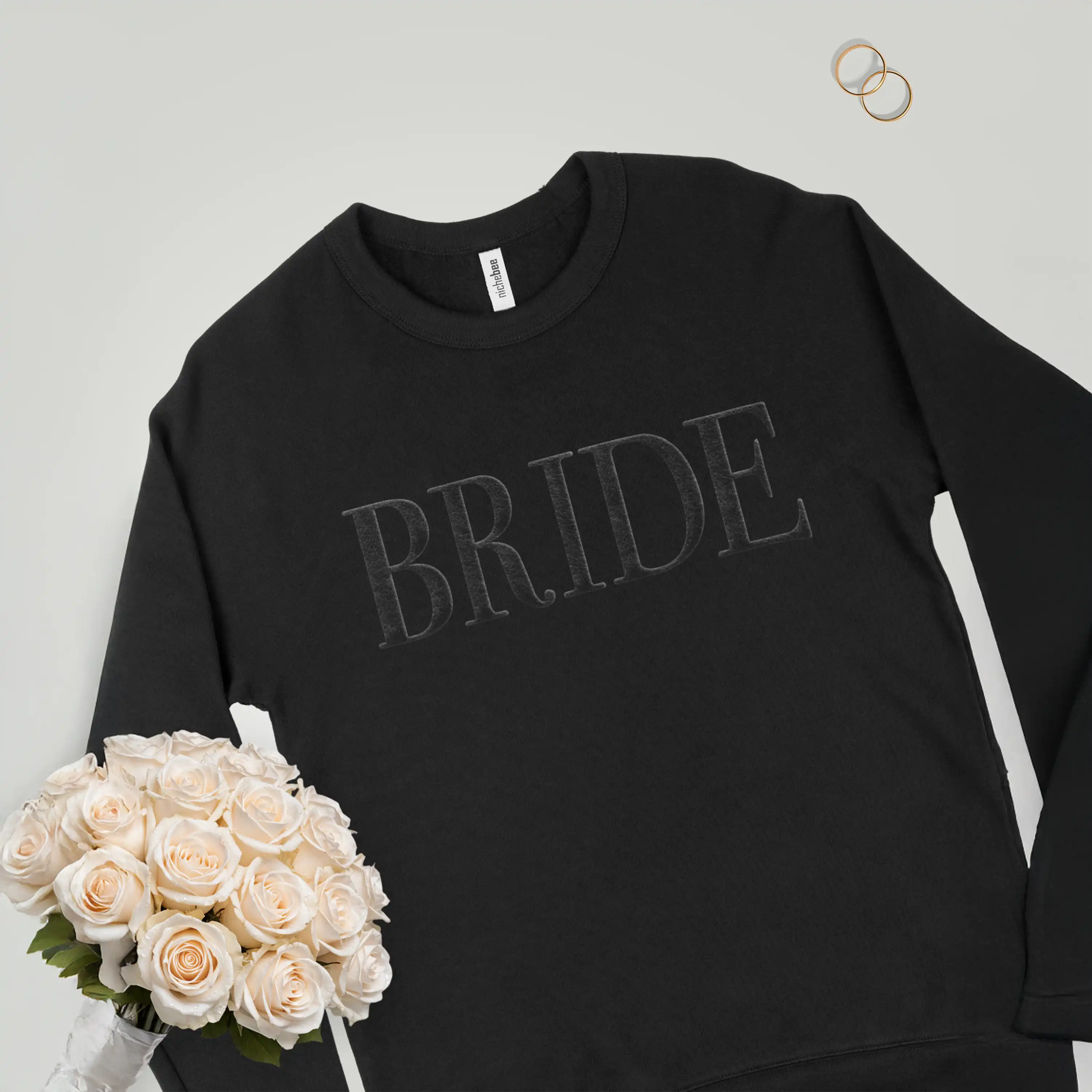 Black sweatshirt with 'BRIDE' text, bouquet of flowers, and wedding rings on a light gray background