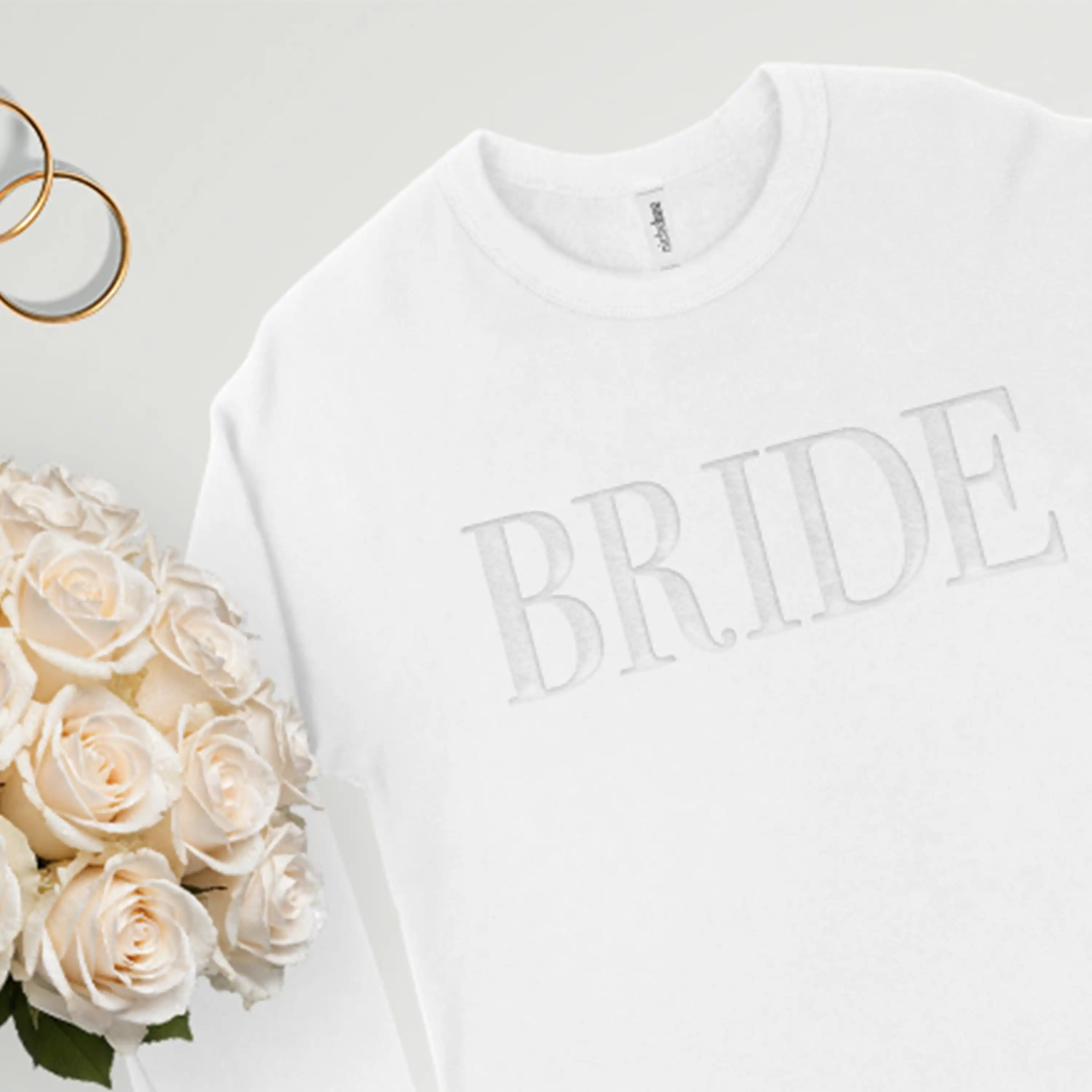 White sweatshirt with 'BRIDE' text on a light background with flowers and rings.