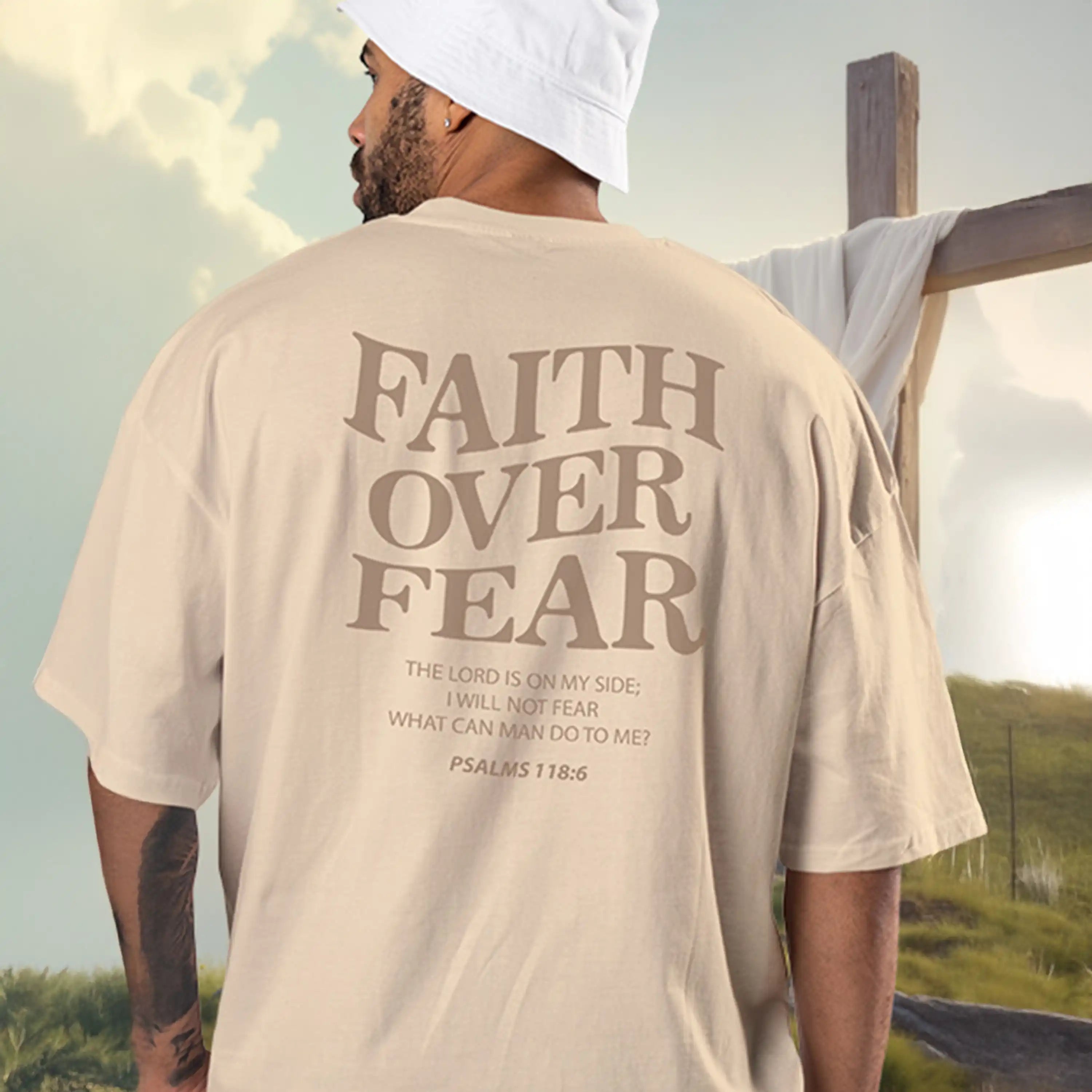 Person wearing a beige t-shirt with 'Faith Over Fear' text, standing near a cross.